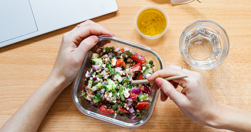 A representation of a woman's hands putting a fork into a salad with a laptop beside her
