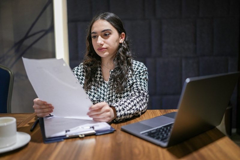 Woman reviewing documents at a desk with a laptop and coffee cup, looking serious and focused.