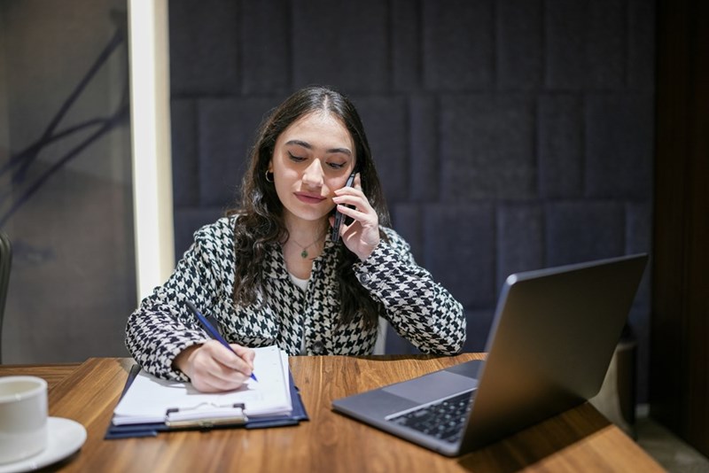 Woman on the phone writing on a clipboard at a desk beside a laptop and coffee cup.