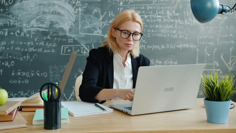 Focused educator using a laptop while organizing notes and materials in a modern classroom setting.