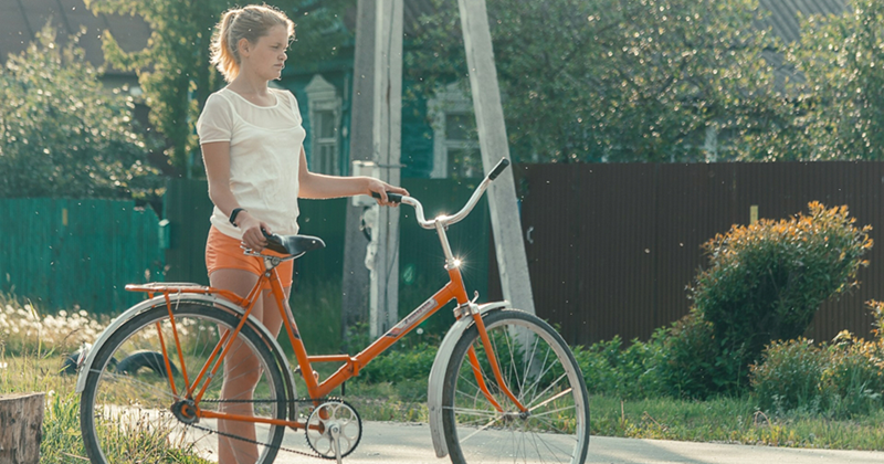 A representation of a teenage girl standing outside, holding an orange bike