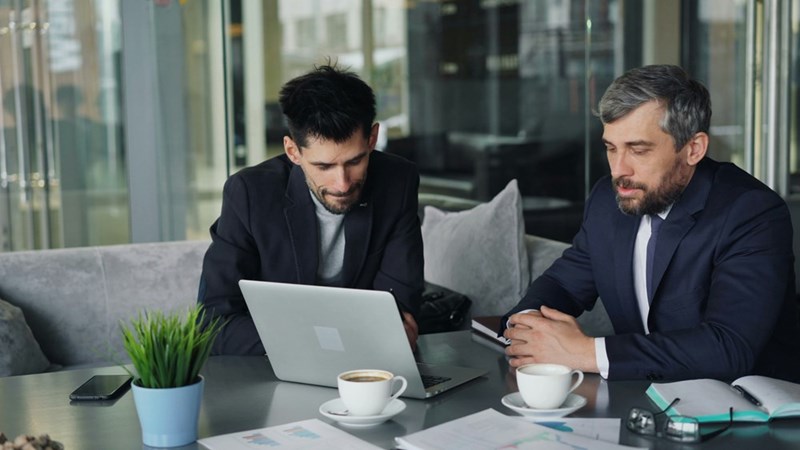 Two men (models) sitting at a table, looking at a laptop