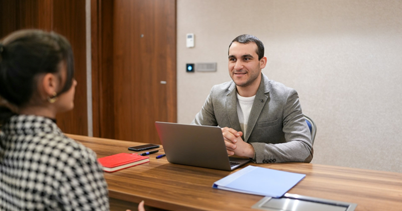 Two models representing a male hiring manager with a laptop, and a female job applicant sitting across the desk from him