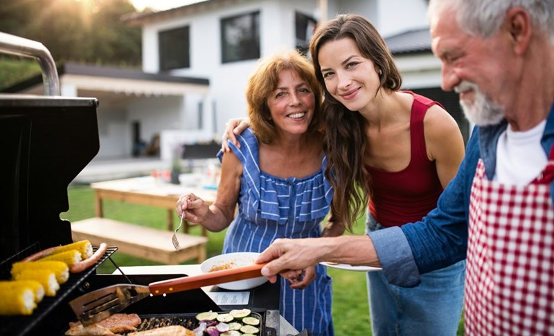 Aunt and her nephew's GF snap a photo together while another family member works the grill, in a representation by models.