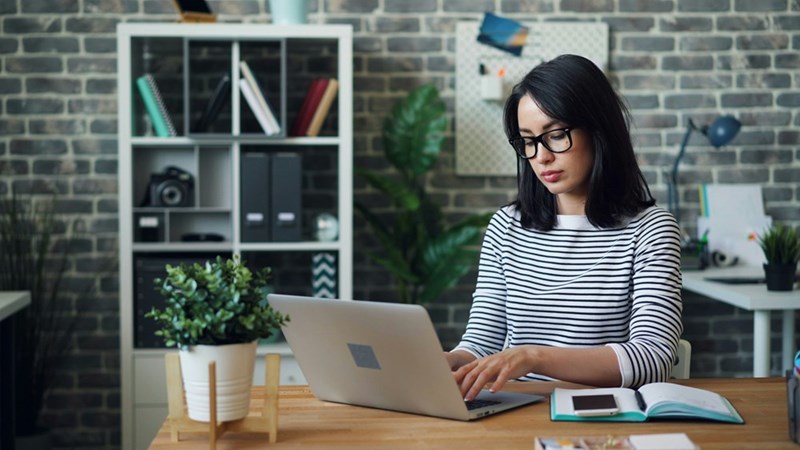 Woman in glasses working on a laptop at an office desk with a notebook and phone beside her.