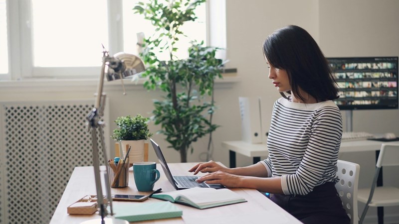 Woman working on a laptop at a desk with a notebook, mug, and plants nearby.