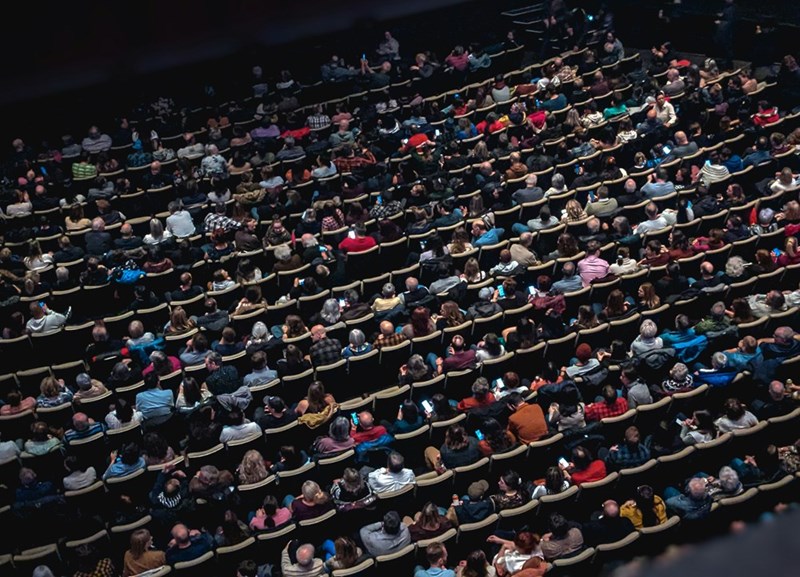 Crowded theater auditorium with rows of seated audience members viewed from above.