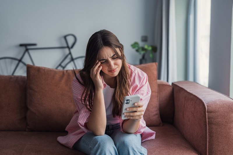 Young woman sitting on a couch looking stressed while holding a phone in a bright living room.