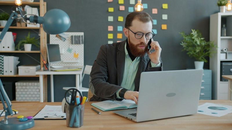 A businessman multitasks between a phone call and computer work during a busy day at the office.