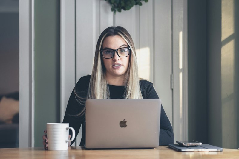 Woman in glasses sitting at a laptop with a mug and phone nearby, looking mildly amused.