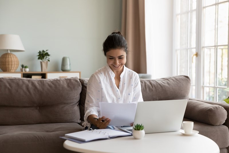 South Asian woman smiling at paperwork beside a laptop while sitting on a couch in a bright living room.