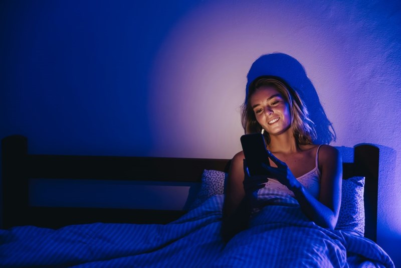 Woman smiling at her phone while sitting up in bed under blue light, looking pleased and relieved.
