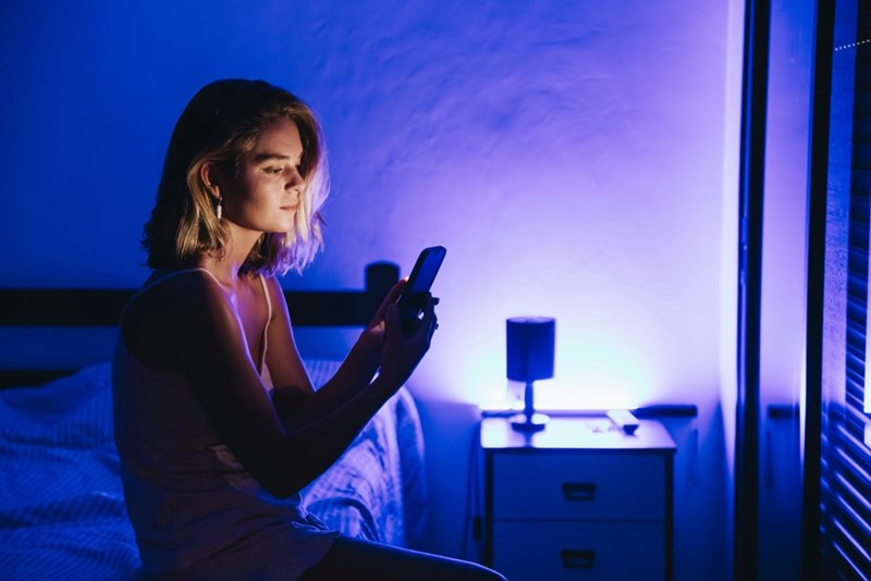 Woman sitting beside a bed in blue light, staring suspiciously at her phone near a nightstand.