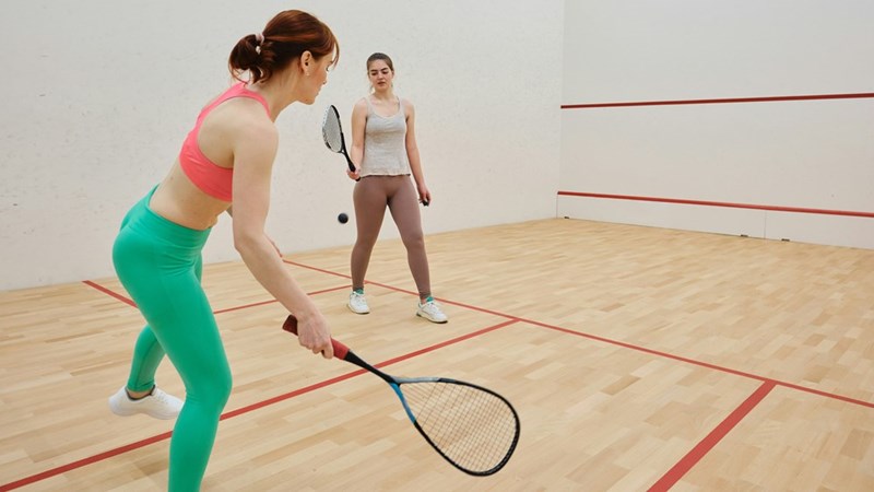 Two women play squash, hitting the ball back and forth in a gym. Subjects are models.
