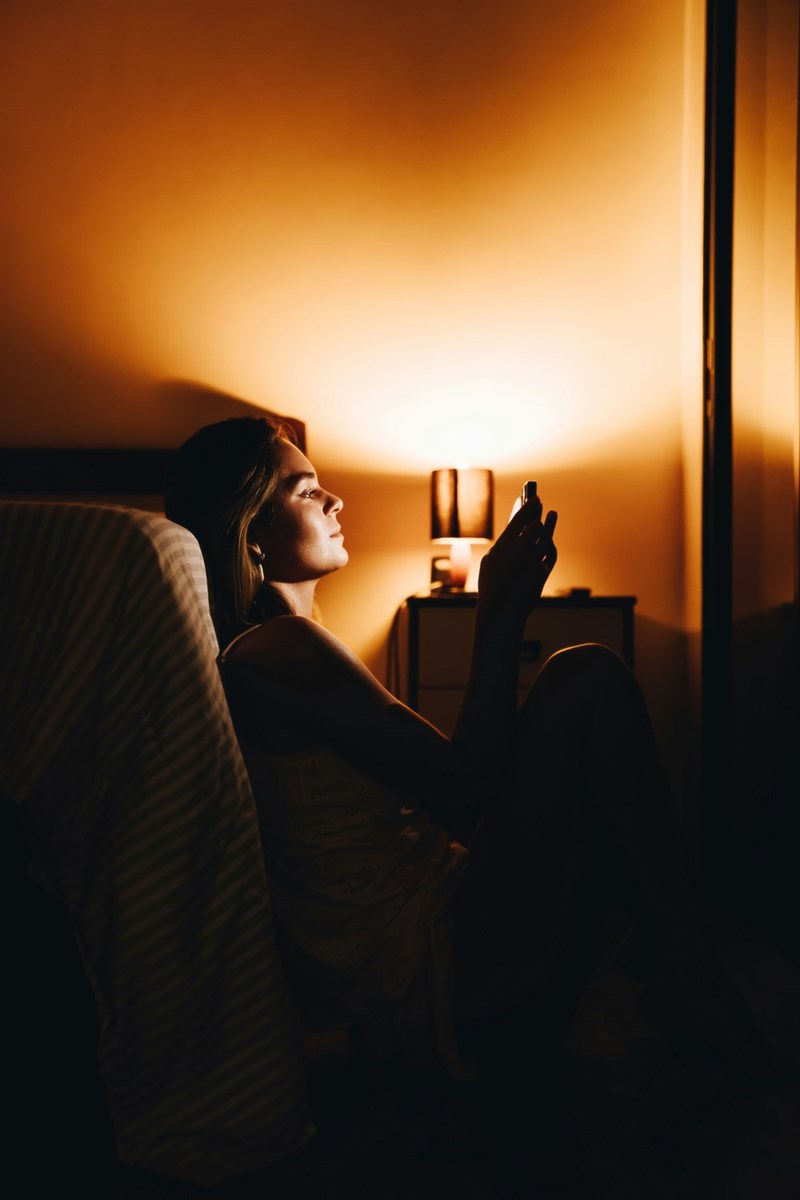 Woman sitting on the floor beside a bed at night, looking worried while reading her phone in warm lamp light.