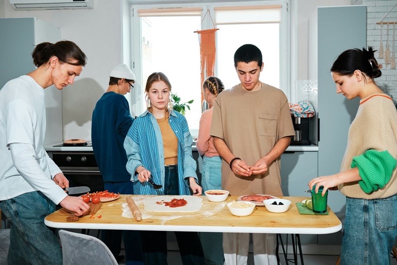 A photograph depicting four friends chopping food in the kitchen while other friends socialize in the background.