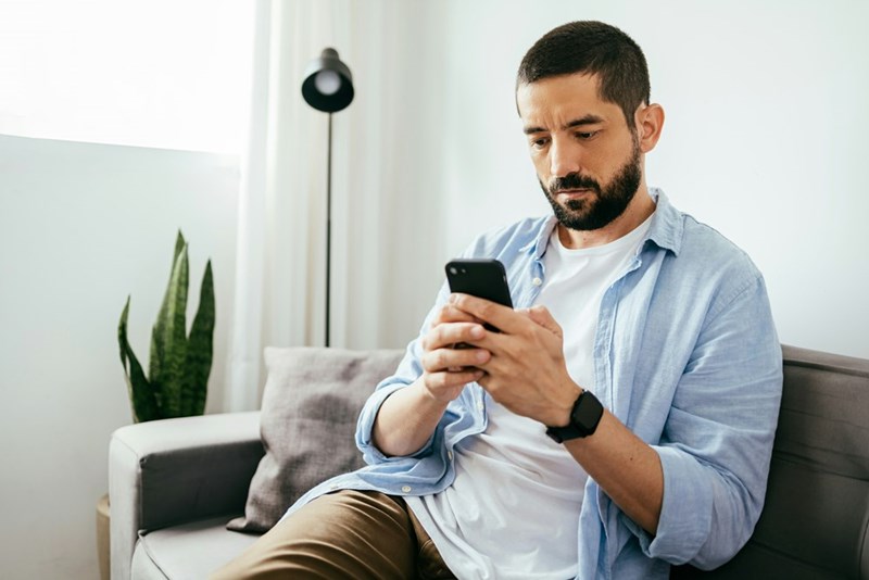 Man with beard sitting in a white couch using his phone.