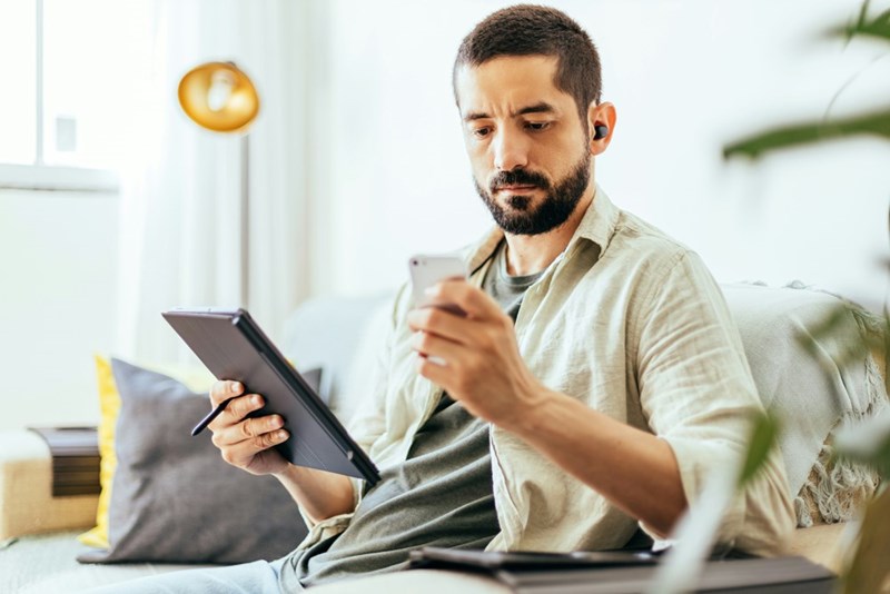 Man with beard holding an ipad on one hand and a phone on another sitting on his couch.