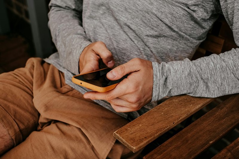 A mans body sitting crossed leg on a bench holding his phone and typing.