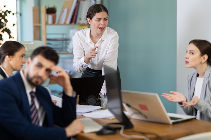 Coworkers arguing around an office table with laptops and papers during a tense workplace meeting.