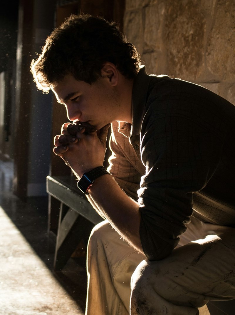 Man sitting alone with clasped hands, looking troubled in dramatic sunlight.