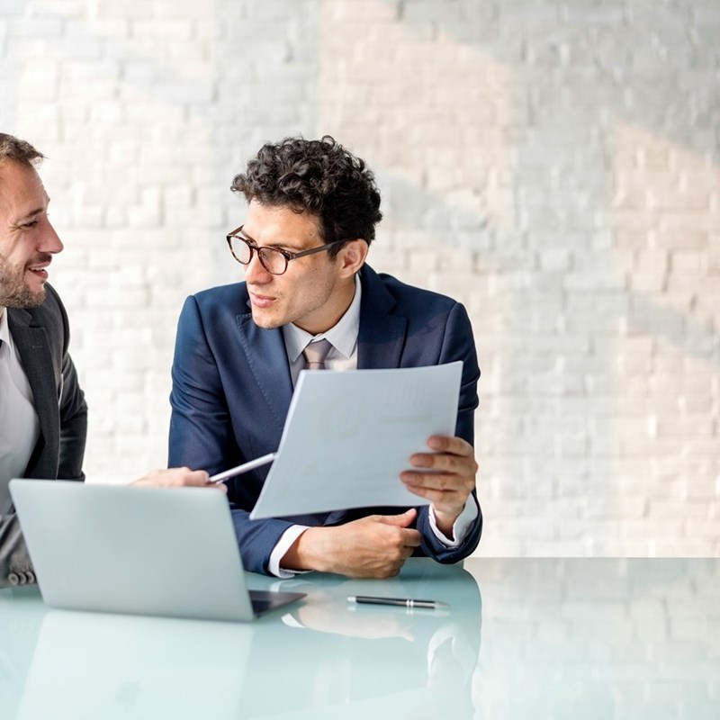 Two men wearing a formal suit sitting down discussing paperwork.