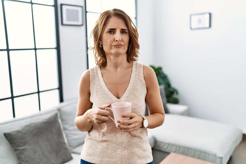 Woman holding a mug in a living room, looking frustrated and skeptical.