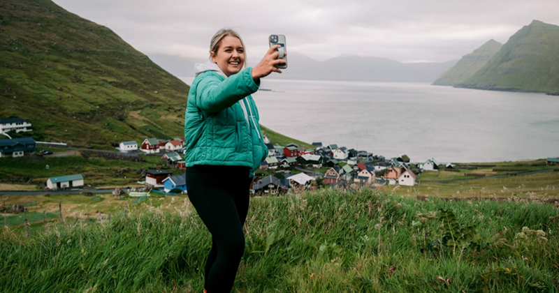 A representation of a woman smiling as she takes a selfie with grassy hills and the sea in the background