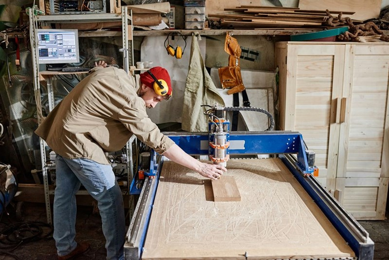 Man operating a woodworking machine in a busy workshop with tools, lumber, and cabinets around him.