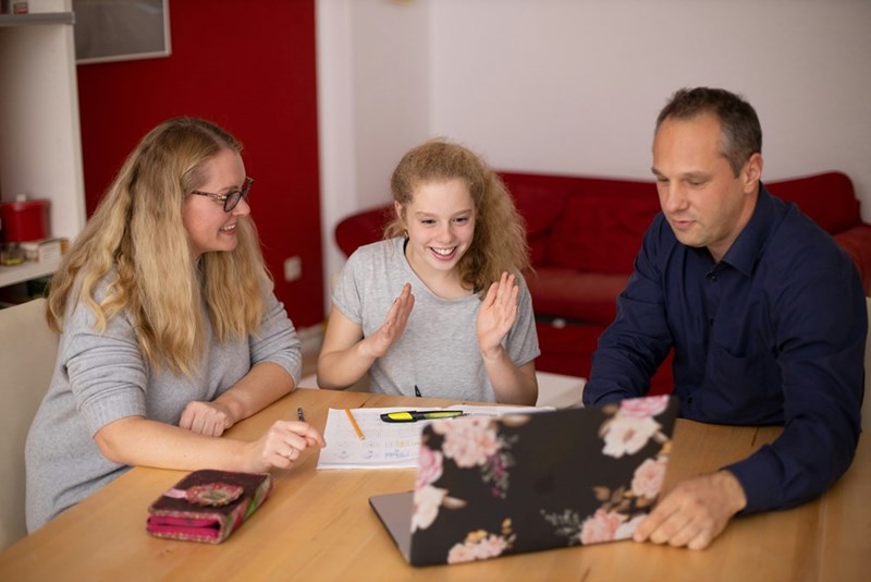 A family looking at a laptop on the table in front of them as the daughter excitedly responds to what they are seeing on the screen