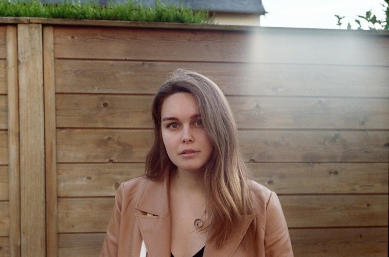 Woman standing outdoors by a wooden fence near a home, looking serious and resolved.
