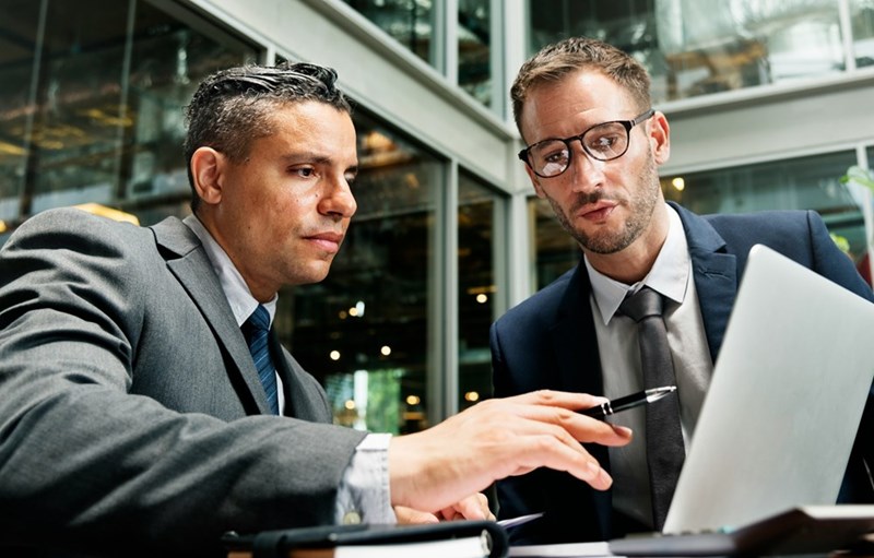 Two professionally dressed businessmen sit in a lobby and go over paperwork together (models).