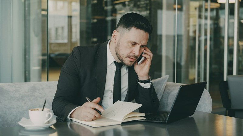 A professionally dressed businessman sits at a cafe table drinking a coffee, writing in a notebook, and having a discussion on his cellphone (model).