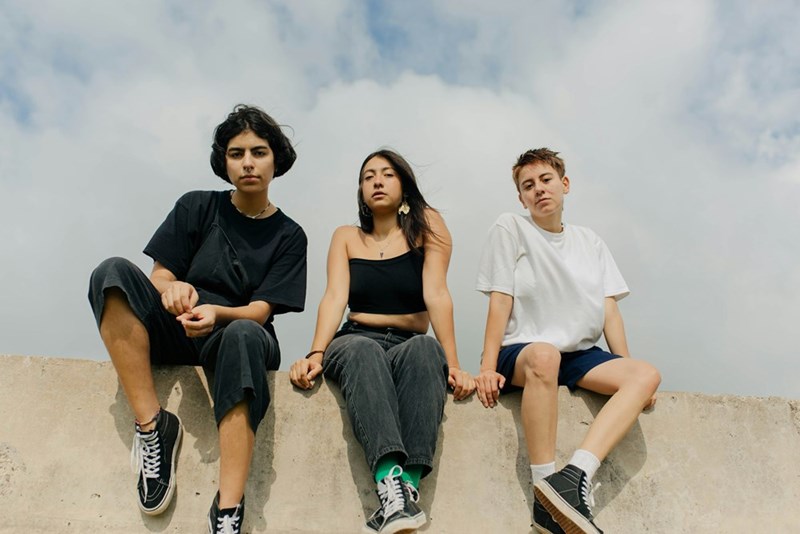 Three young people sit together on a concrete wall under an open sky.