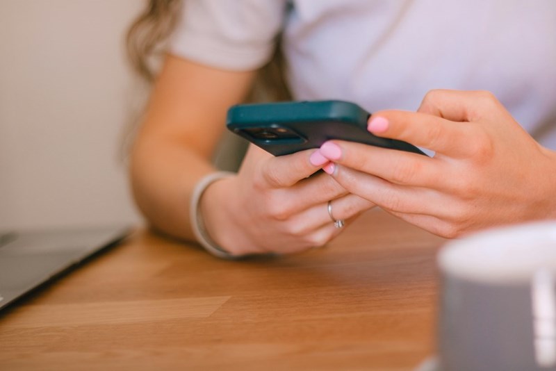 A person holds a smartphone while sitting at a desk with a laptop nearby.