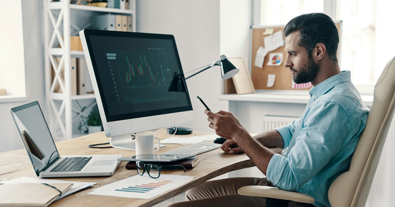 A representation of an employee sitting in front of a computer screen with a graph on it and looking at his phone