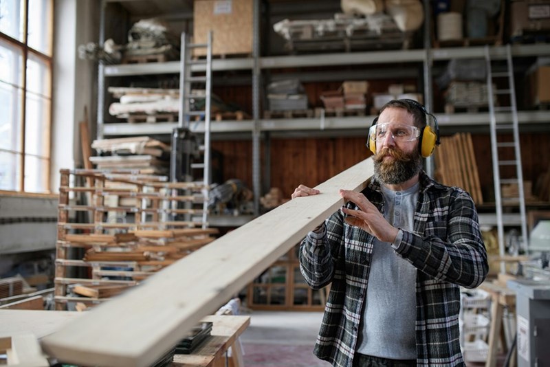 a mature male carpenter with hearing protectors carrying wooden board indoors in carpentry workshop small business concept