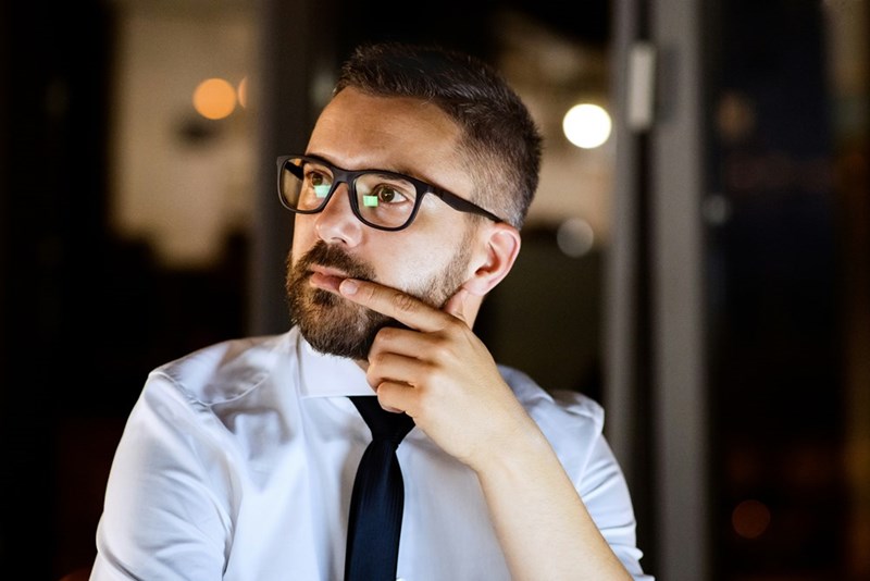 Man in glasses and a tie looking thoughtful at night with his hand on his chin.