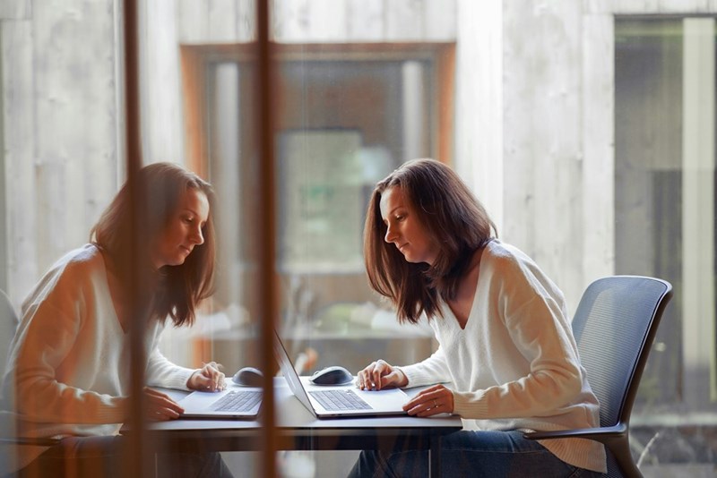 Woman working alone at a laptop in a bright office, seen through glass with her reflection beside her.