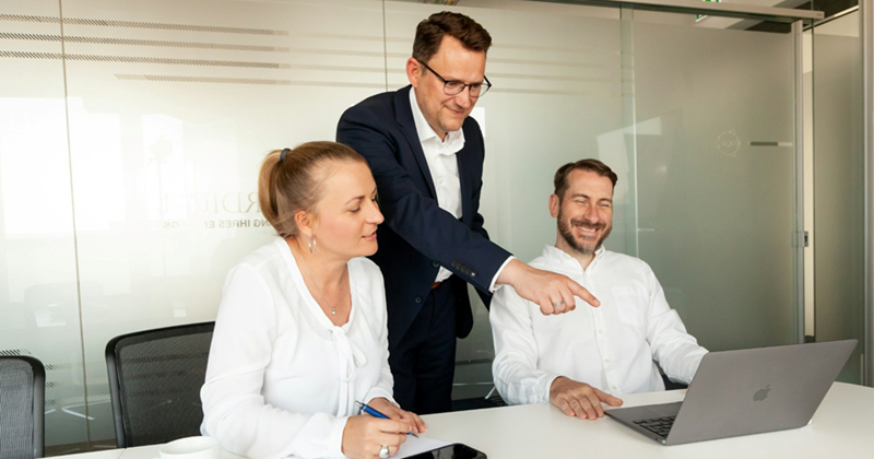 A representation of two employees sitting with a laptop at a meeting table, with the boss standing behind them and pointing at the screen