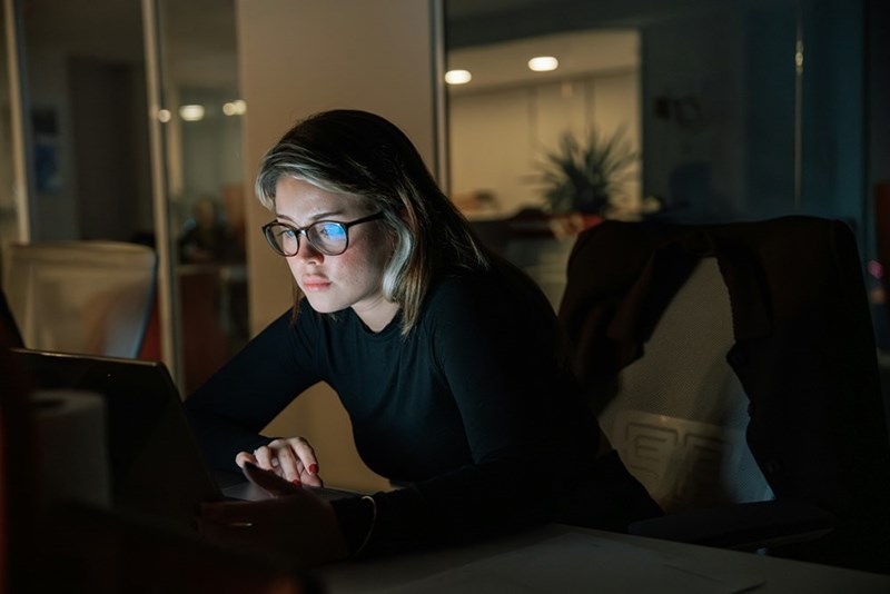 Young woman in glasses working late on a laptop in a dim room, looking serious and focused.
