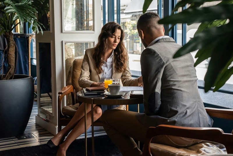 Woman and man seated at a small table in a tense work meeting with papers, coffee, and a glass of orange juice.