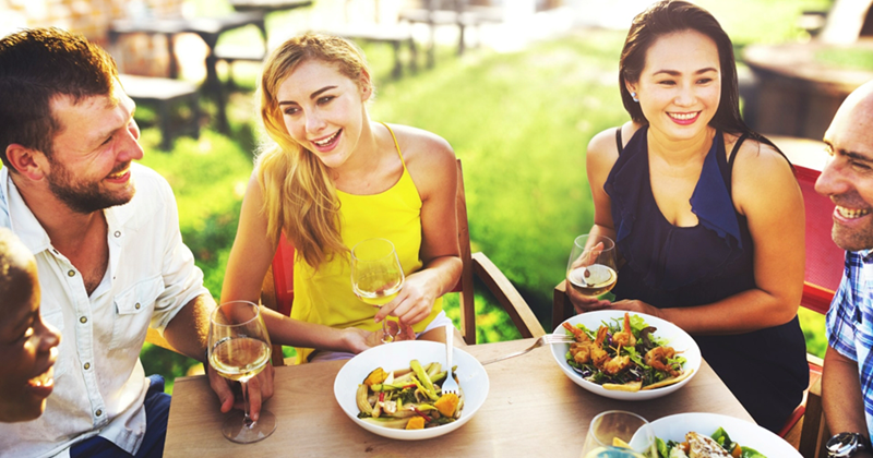 A representation of friends and family members eating and drinking at an outside table at a restaurant
