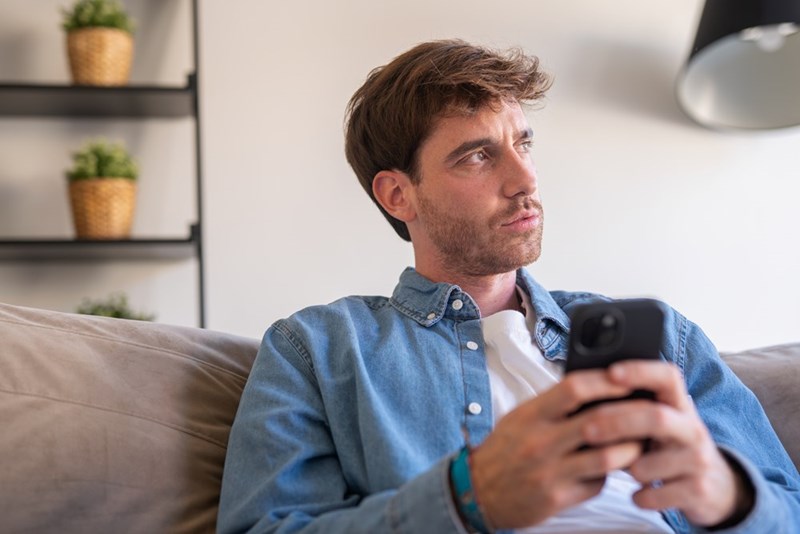 Man in a denim shirt sitting on a couch, holding his phone and looking off to the side.