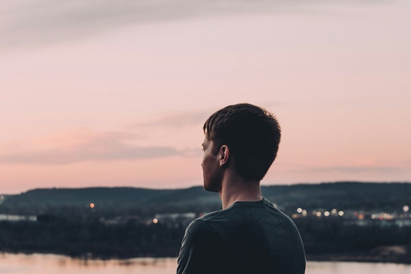 Young man seen from behind, looking over a river and hills beneath a pink sunset sky.