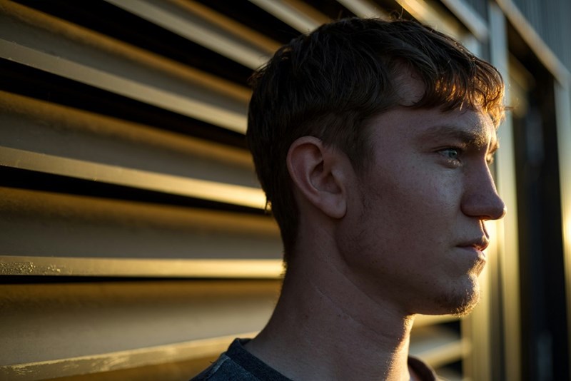 Young man in profile standing beside metal siding in warm sunlight, looking into the distance.
