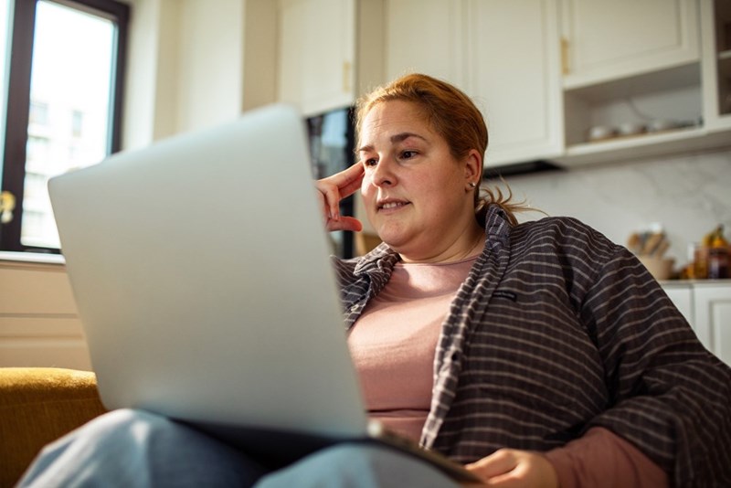 Woman sitting with a laptop in a bright kitchen, resting her hand near her temple and looking concerned.