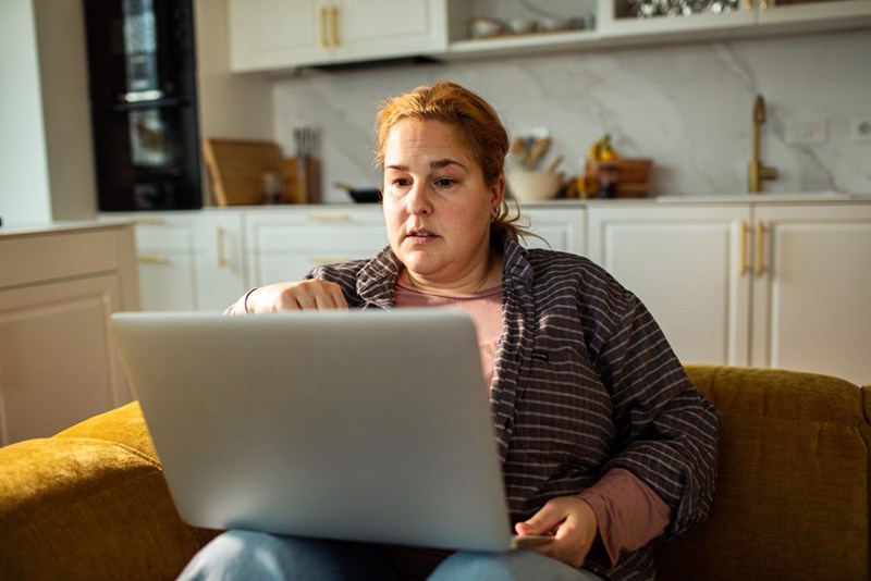 Woman sitting on a mustard couch with a laptop in a bright kitchen, looking concerned
