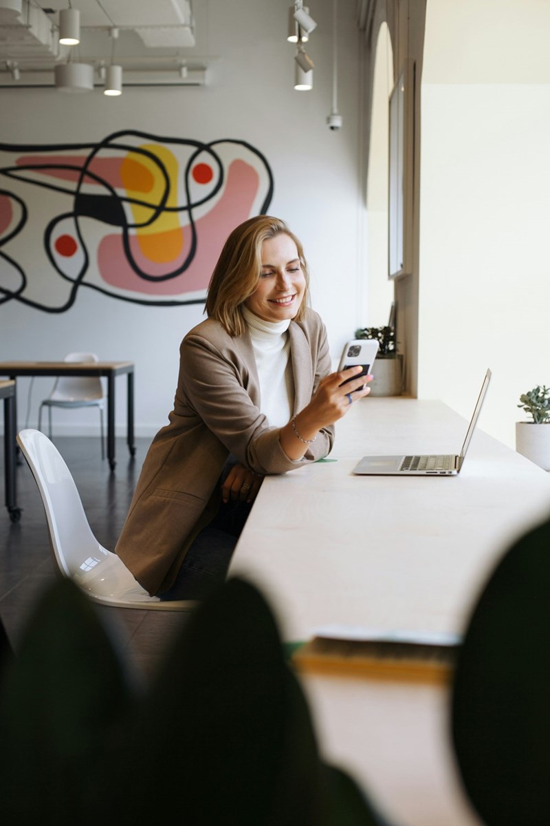 Woman smiling at her phone beside a laptop at a long table in a modern workspace with colorful wall art.