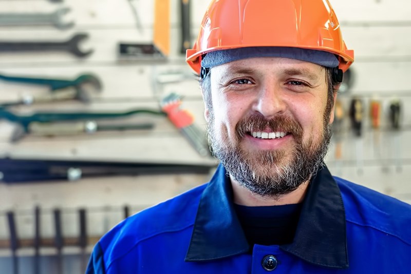 Smiling man in a hard hat and blue work jacket standing in a workshop with tools behind him